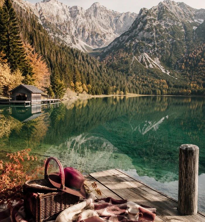 Alpensee mit Picknickkorb, Hut und Bergspiegelung